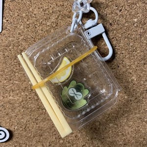 Op de afbeelding: Een sleutelhanger van een doorzichtige plastic container met een citroenpartje en een groen kikkerachtig ontwerp aan de binnenkant. De container is vastgezet met een gele elastiek en wordt geleverd met een paar beige eetstokjes. Een witte metalen sluiting en een stervormige bedel zijn bevestigd.