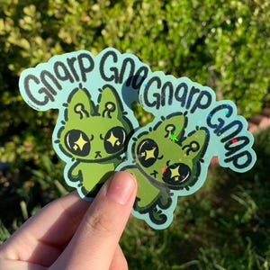 Pode incluir: Dois adesivos holográficos verdes com um personagem alienígena de desenho animado e o texto "Gnar Gnar Gnar Gnar" em preto.