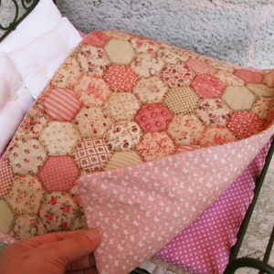 Op de afbeelding: Een poppenbed met een wit metalen frame en een roze en witte patchworkdeken. De deken is gemaakt van zeshoekige stofstukken met bloemen- en geometrische patronen.
