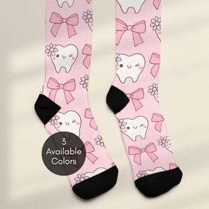 May include: A pair of light pink socks featuring a repeating pattern of white cartoon teeth with winking faces, light purple flowers, and pink bows. The socks have black heels and black toe sections. A circular black badge displays the text 3 Available Colors.