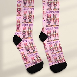May include: A pair of pink socks featuring a repeating pattern of watercolor-style nutcracker soldiers. The socks have black heels and toes, with the words "What's Cracking?" and "What's Crac" printed on them.