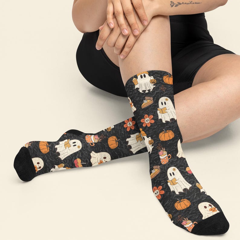 Puede incluir: Un par de calcetines negros con un patr&oacute;n repetitivo de fantasmas blancos, calabazas naranjas, flores naranjas con centros de calavera y tazas de bebidas calientes. Los calcetines tienen talones y puntas de color negro s&oacute;lido.