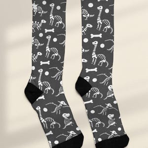 May include: A pair of knee-high socks with a dark gray background and a repeating pattern of white dinosaur skeletons, bones, and circles. The socks have black heels and toes.