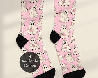 May include: A pair of light pink crew socks featuring an all-over pattern of white ghosts dressed as nurses, complete with medical caps, stethoscopes, and first aid kits. The socks have black heels and toes, with a circular badge stating 4 Available Colors.