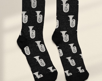 Puede incluir: Un par de calcetines negros con un patrón repetido de ilustraciones de tuba blancas. Los calcetines tienen talones y punteras negras, y el diseño de la tuba está espaciado uniformemente. Un accesorio divertido para los amantes de la música.