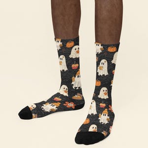 Puede incluir: Un par de calcetines de corte medio con fondo gris oscuro y un patr&oacute;n de fantasmas blancos, calabazas naranjas, trozos de tarta, manzanas de caramelo y flores naranjas. Los calcetines tienen las punteras y los talones en color negro.