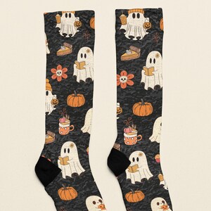 Puede incluir: Un par de calcetines negros con un patr&oacute;n repetitivo de fantasmas blancos, calabazas naranjas, porciones de pastel de calabaza, tazas naranjas con crema batida y flores naranjas con centros en forma de calavera.