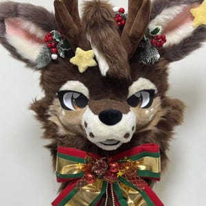 May include: A brown furry deer head with large ears, antlers, and a red and green bow with gold ribbon and jingle bells. The deer head is decorated with red berries, green leaves, and gold stars.