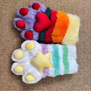 Puede incluir: Un par de guantes de pata de felpa coloridos. Un guante presenta piel morada con almohadillas rojas y un corazón rojo. El otro guante es blanco con almohadillas amarillas y una estrella amarilla, con rayas pastel.