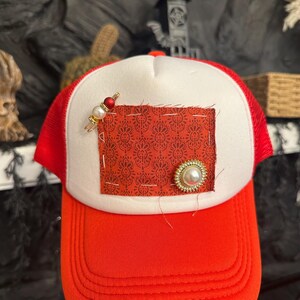 Pu&ograve; includere: Cappellino da camionista rosso e bianco con retro in rete rossa e visiera curva. La parte anteriore presenta una toppa fantasia rossa con un abbellimento di perle e oro e una spilla decorativa con perline rosse e bianche.