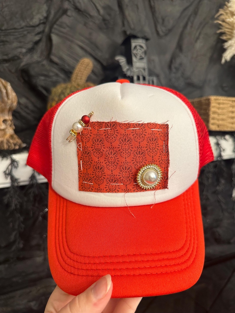 Pu&ograve; includere: Cappellino da camionista rosso e bianco con retro in rete rossa e visiera curva. La parte anteriore presenta una toppa fantasia rossa con un abbellimento di perle e oro e una spilla decorativa con perline rosse e bianche.