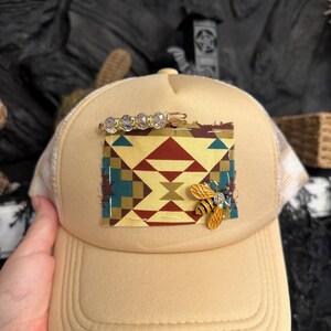 Pu&ograve; includere: Cappellino da camionista beige con retro in rete color crema. La parte anteriore presenta una toppa con un disegno geometrico in rosso, turchese e oro. Una spilla a forma di ape dorata e una barretta di perline di cristallo adornano il cappello.