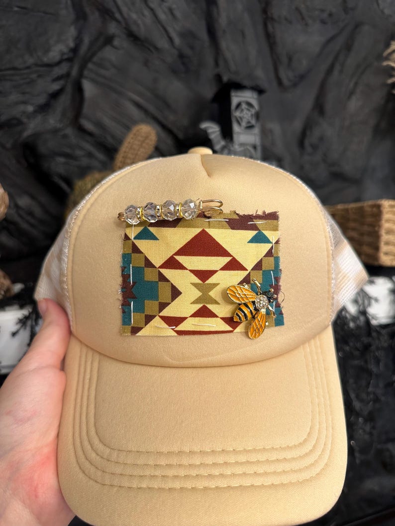 Pu&ograve; includere: Cappellino da camionista beige con retro in rete color crema. La parte anteriore presenta una toppa con un disegno geometrico in rosso, turchese e oro. Una spilla a forma di ape dorata e una barretta di perline di cristallo adornano il cappello.
