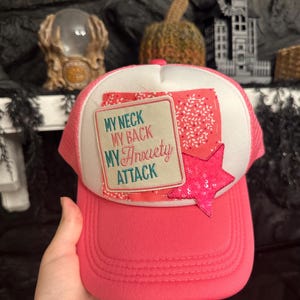 Pu&ograve; includere: Cappellino da camionista rosa con pannello frontale bianco con la scritta "MY NECK MY BACK MY Anxiety ATTACK". &Egrave; attaccata una stella rosa con paillettes. Il cappellino ha una visiera curva e un retro in rete.
