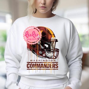 May include: White sweatshirt featuring a football helmet design in burgundy, gold, and brown. The words "WASHINGTON COMMANDERS" are printed below the helmet in a matching color scheme. The sweatshirt has long sleeves.
