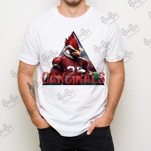 Pu&ograve; includere: Maglietta bianca con una grafica rossa, bianca e nera di un cardinale dei cartoni animati che indossa una divisa da football e la parola "CARDINALS" in un font rosso, bianco e nero.