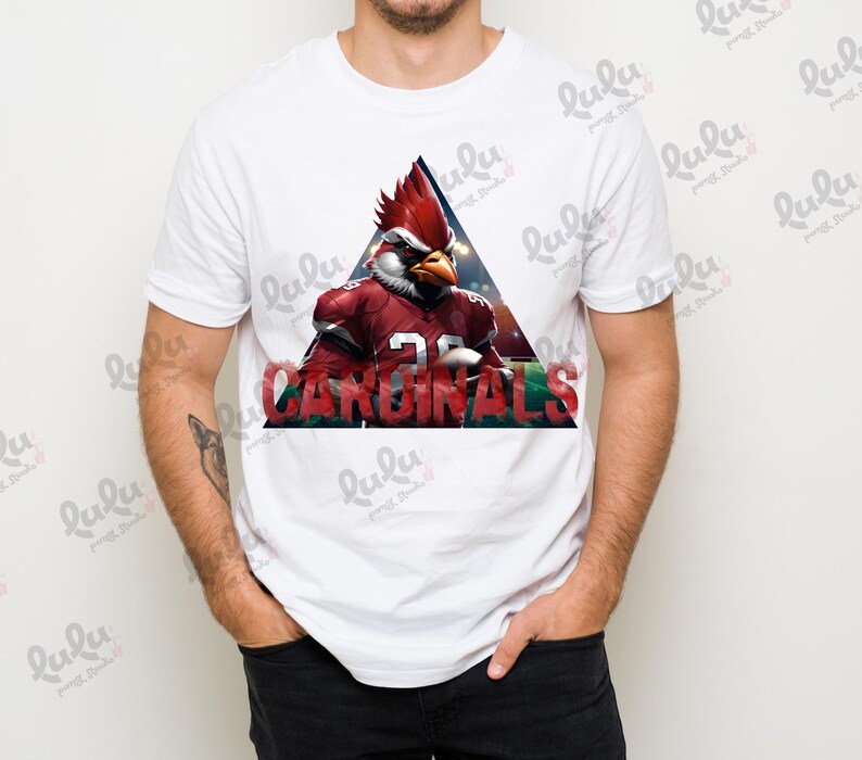Pu&ograve; includere: Maglietta bianca con una grafica rossa, bianca e nera di un cardinale dei cartoni animati che indossa una divisa da football e la parola "CARDINALS" in un font rosso, bianco e nero.