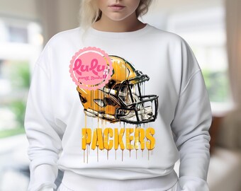 May include: White sweatshirt featuring a gold and green football helmet graphic with the word "PACKERS" in yellow. The design has a dripping paint effect. The sweatshirt has long sleeves and a crew neck.