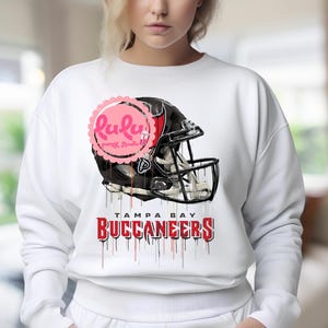 May include: White sweatshirt featuring a Tampa Bay Buccaneers football helmet graphic. The helmet is black with red accents and a silver facemask. The team name is printed in red with a dripping effect. A pink logo is visible.