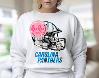 May include: White sweatshirt with a Carolina Panthers football helmet design. The helmet is in blue and grey, with the team name in blue below. A pink logo is visible on the upper left.
