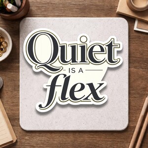 May include: A square, off-white sticker with the words "Quiet is a flex" in a stylized black font with a gold outline. The sticker is on a light gray surface, with office supplies in the background.