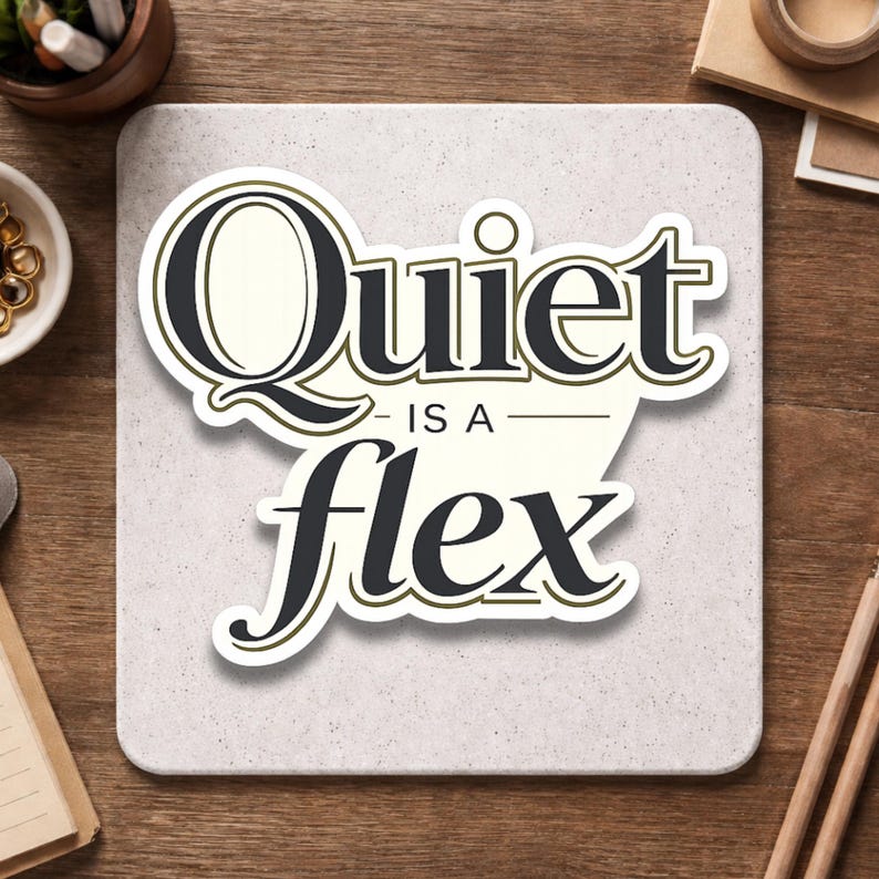 May include: A square, off-white sticker with the words "Quiet is a flex" in a stylized black font with a gold outline. The sticker is on a light gray surface, with office supplies in the background.