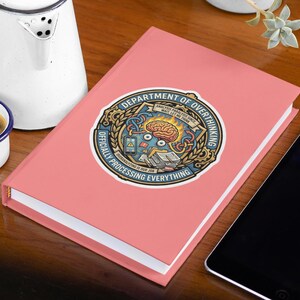 May include: A pink journal with a sticker that reads "DEPARTMENT OF OVERTHINKING OFFICIALLY PROCESSING EVERYTHING." A small white mug of coffee and a white kettle are next to the journal. A black tablet is on the right.