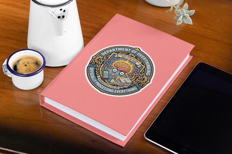 May include: A pink journal with a sticker that reads "DEPARTMENT OF OVERTHINKING OFFICIALLY PROCESSING EVERYTHING." A small white mug of coffee and a white kettle are next to the journal. A black tablet is on the right.