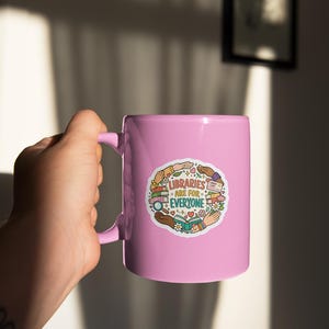 May include: A light pink ceramic mug with a handle, featuring a colorful sticker that reads "LIBRARIES ARE FOR EVERYONE." The sticker includes illustrations of books, hands, and flowers. The mug is held by a person.