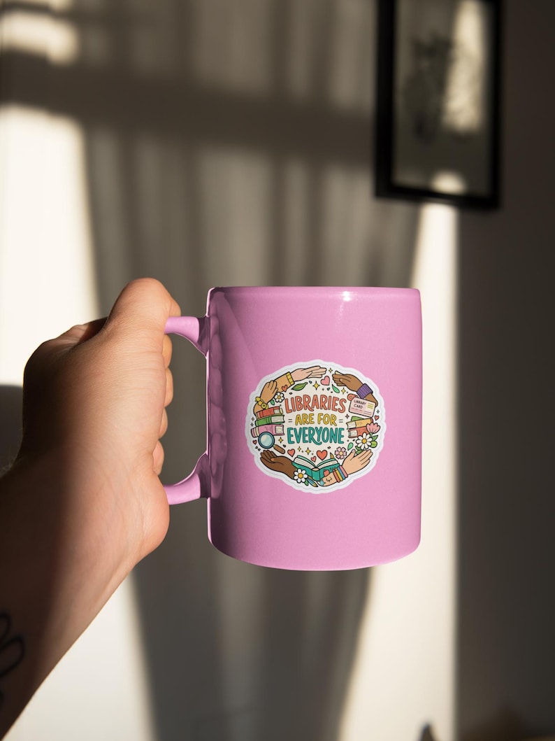 May include: A light pink ceramic mug with a handle, featuring a colorful sticker that reads "LIBRARIES ARE FOR EVERYONE." The sticker includes illustrations of books, hands, and flowers. The mug is held by a person.