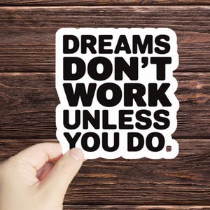 May include: White sticker with black text that reads "Dreams don't work unless you do."