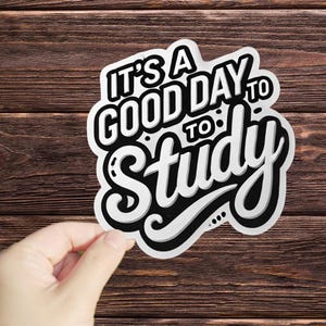 May include: A white sticker with black lettering that reads "It's a good day to study". The sticker is on a brown wooden background.