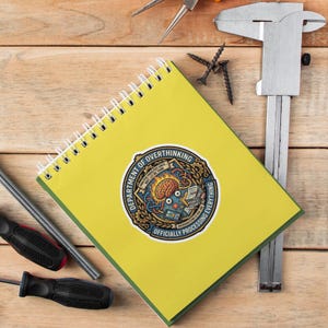 May include: A yellow spiral notebook with a sticker that reads "DEPARTMENT OF OVERTHINKING OFFICIALLY PROCESSING EVERYTHING." The sticker features a brain graphic and other elements. The notebook is on a wooden surface with tools.