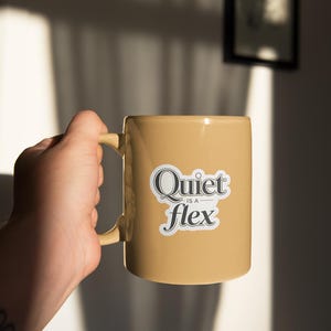 May include: A tan ceramic mug with a curved handle. The mug features the text "Quiet is a flex" in a black and white sticker. The mug is held by a person's hand, with a tattoo visible on the wrist.