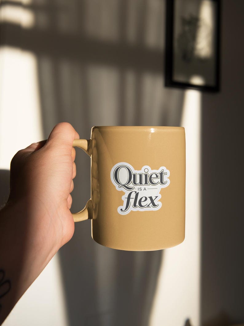May include: A tan ceramic mug with a curved handle. The mug features the text "Quiet is a flex" in a black and white sticker. The mug is held by a person's hand, with a tattoo visible on the wrist.