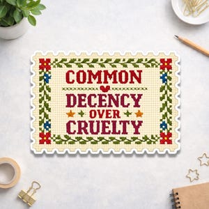 May include: A sticker with a cross-stitch design. The text reads "COMMON DECENCY OVER CRUELTY" in burgundy, with a heart and stars. The border is a cream color with a floral design in green, red, and blue.