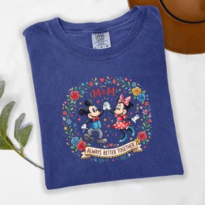 Puede incluir: Una camiseta azul Comfort Colors con Mickey y Minnie Mouse. El dise&ntilde;o incluye una corona floral, las letras "M&M" y la frase "ALWAYS BETTER TOGETHER". Los personajes est&aacute;n representados con sus atuendos cl&aacute;sicos.