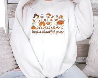May include: White sweatshirt featuring a Thanksgiving-themed graphic. The design includes five cartoon geese wearing hats and holding fall-themed items. The text "Just a thankful goose" is printed below the geese in brown script.