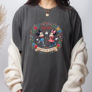 Puede incluir: Camiseta gris oscuro con Mickey y Minnie Mouse en una corona floral. El dise&ntilde;o incluye el texto "MOM" y "Always Better Together" en una pancarta. Mickey lleva un chaleco azul y Minnie un vestido de lunares rojos.