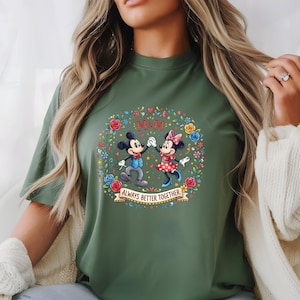Puede incluir: Camiseta verde oliva con Mickey y Minnie Mouse tomados de la mano, rodeados por una corona floral. Tambi&eacute;n se muestran los textos "M&M" y "Always Better Together". El dise&ntilde;o est&aacute; centrado en la camiseta.