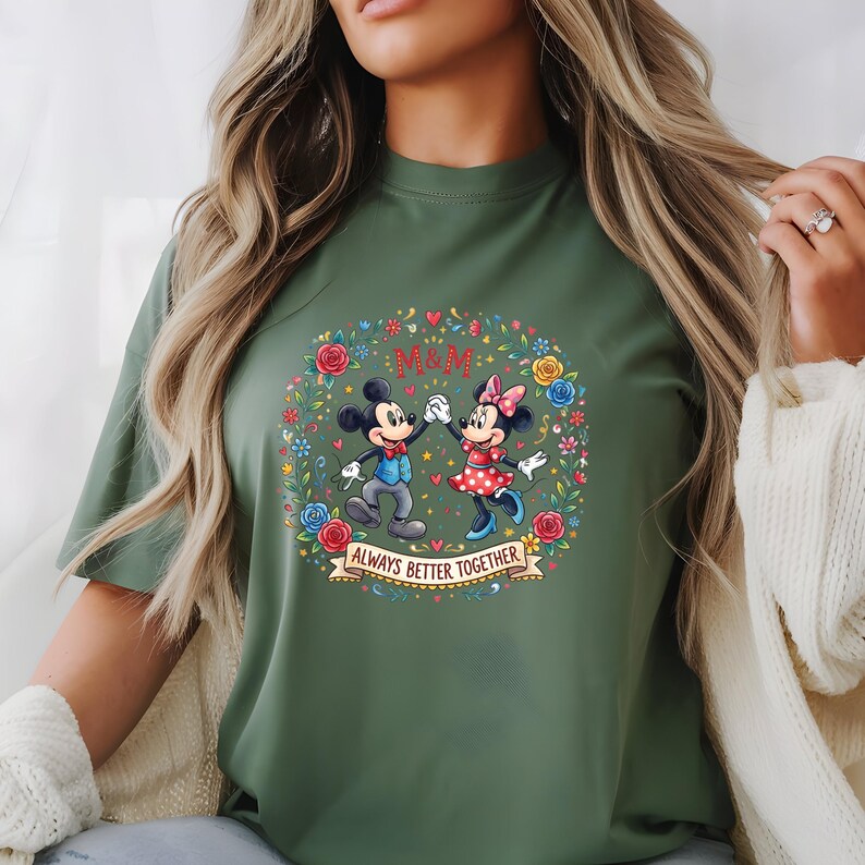 Puede incluir: Camiseta verde oliva con Mickey y Minnie Mouse tomados de la mano, rodeados por una corona floral. Tambi&eacute;n se muestran los textos "M&M" y "Always Better Together". El dise&ntilde;o est&aacute; centrado en la camiseta.