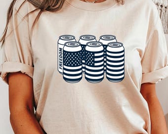 May include: A beige t-shirt featuring a graphic of six beer cans. Two cans display the American flag design, while the others have a striped pattern. The word "FREEDOM" is printed vertically on one can.