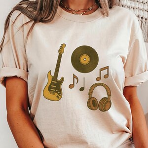 May include: Beige t-shirt featuring a musical theme. The design includes a yellow guitar, a vinyl record, musical notes, and headphones. The graphics are in shades of yellow, brown, and black.