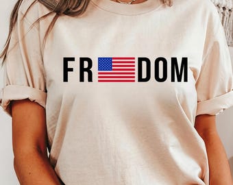 May include: Beige t-shirt with the word "FR DOM" in black block letters. The letters "FR" and "DOM" frame a graphic of the American flag. The shirt has rolled-up sleeves and a relaxed fit.
