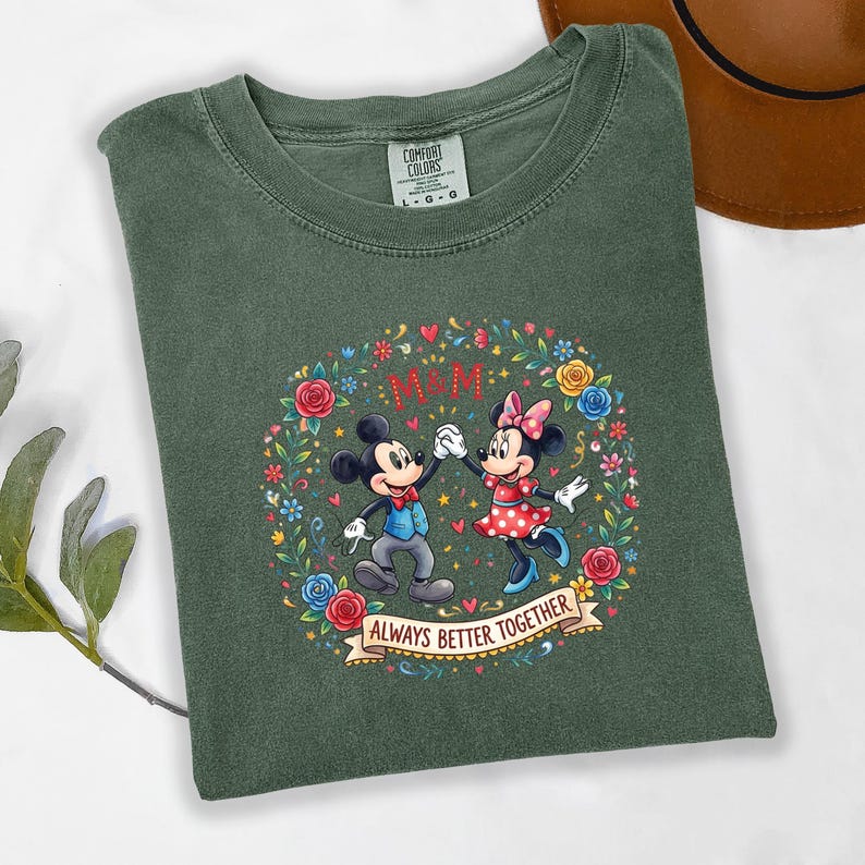 Puede incluir: Camiseta verde con un dise&ntilde;o de Mickey y Minnie Mouse tomados de la mano, rodeados por una corona floral. El dise&ntilde;o incluye el texto "M&M" y "ALWAYS BETTER TOGETHER" en una pancarta. La camiseta es de la marca Comfort Colors.