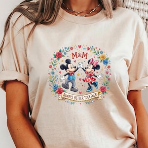 Puede incluir: Camiseta beige con un gr&aacute;fico colorido de Mickey y Minnie Mouse tomados de la mano, rodeados por una corona floral. El texto "M&M" est&aacute; encima de los personajes, y "ALWAYS BETTER TOGETHER" debajo.