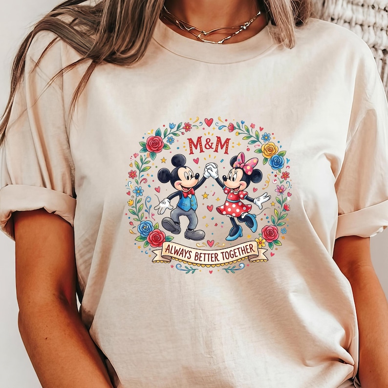 Puede incluir: Camiseta beige con un gr&aacute;fico colorido de Mickey y Minnie Mouse tomados de la mano, rodeados por una corona floral. El texto "M&M" est&aacute; encima de los personajes, y "ALWAYS BETTER TOGETHER" debajo.
