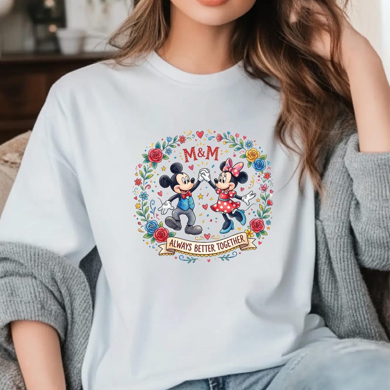 Puede incluir: Camiseta blanca con un gr&aacute;fico colorido de Mickey y Minnie Mouse tomados de la mano, rodeados por una corona floral. Tambi&eacute;n se muestra el texto "M&M" y "ALWAYS BETTER TOGETHER".