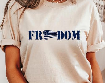 May include: A beige t-shirt with the word "FREEDOM" printed in navy blue. The "E" in "FREEDOM" is replaced with a stylized American flag design. The shirt has short sleeves and a crew neck.