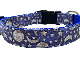 May include: A blue pet collar featuring a repeating pattern of silver-toned moons and stars against a dark blue background. The collar includes a black plastic side-release buckle, a metal adjustment slider, and a silver-toned metal D-ring attachment.